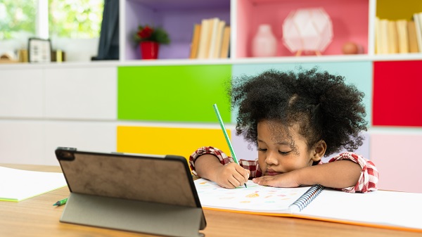 A young girl draws in a notebook while sitting facing an iPad.